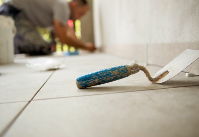 Trowel is on the floor and behind is a worker gluing ceramic til Trowel is on the floor and behind is a worker gluing ceramic tiles.